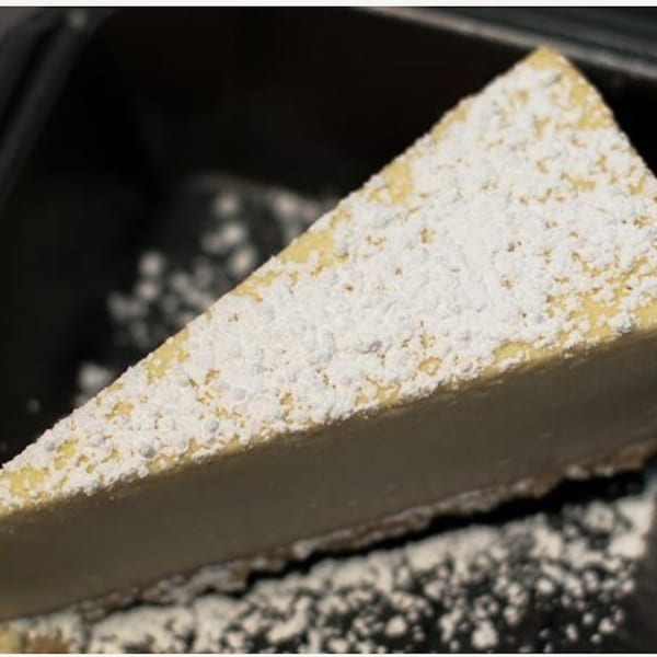 A slice of cheesecake from Pizza Nation, dusted with powdered sugar, on a dark surface.